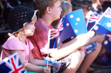 Children in sunshine and holding Australian flgs look outward at a parade